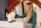 a pregnant person with long blonde hair stroking a white-and-grey cat sitting on a white kitchen counter